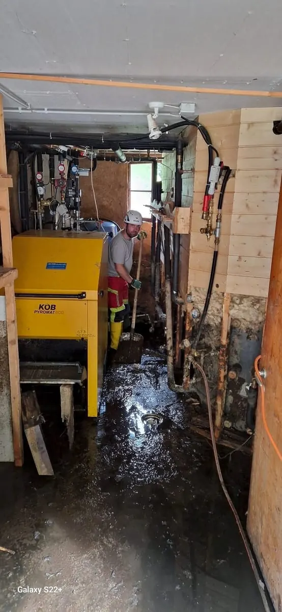 Mann mit Helm und Stiefeln schaufelt Schlamm im Heizungsraum eines überschwemmten Hauses